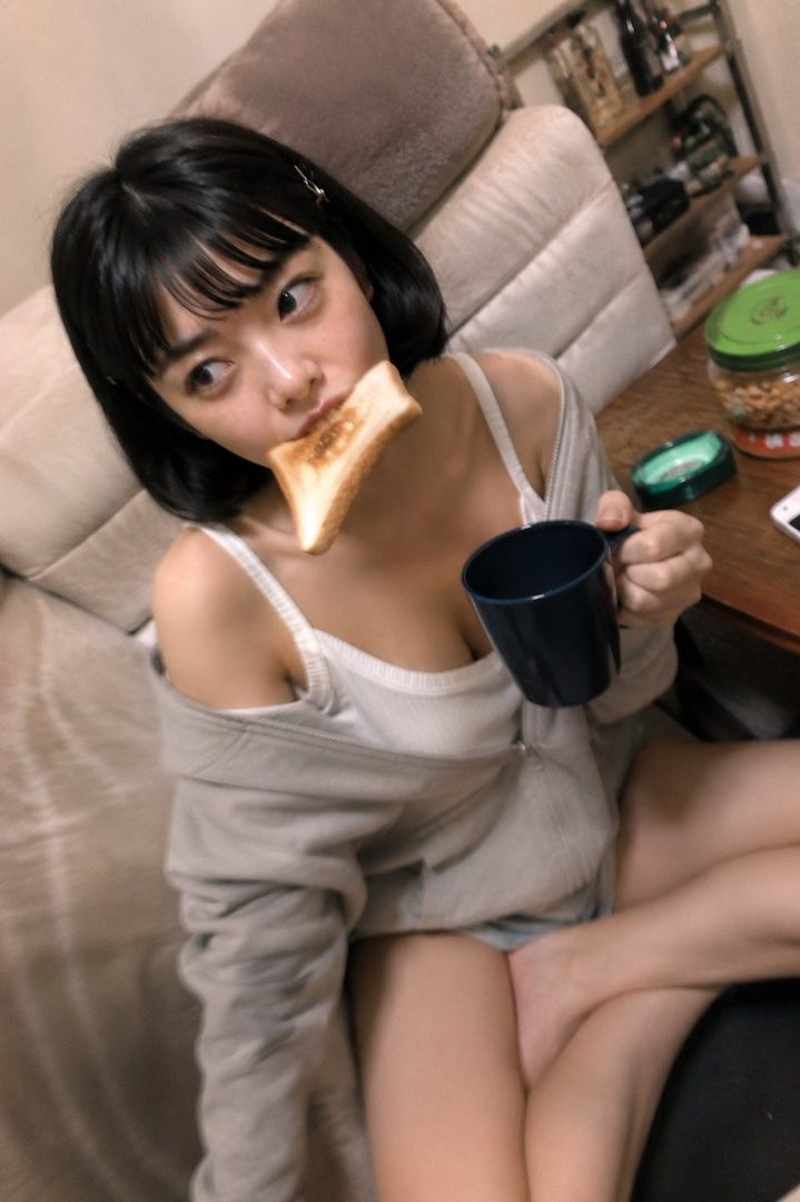 A young woman with short black hair is holding a mug and a piece of toast in her mouth, looking playfully at the camera.