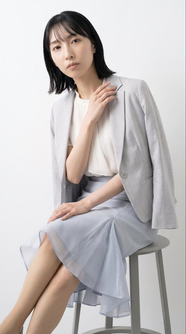 A young East Asian woman with short black hair, wearing a light grey blazer over a white blouse and a light blue skirt, sits on a stool in a studio setting.