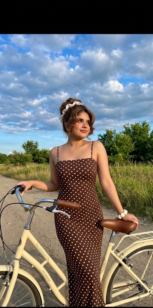 A young woman in a polka-dot dress stands with a bicycle on a dirt road under a cloudy sky.