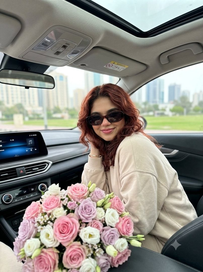 A young woman with wavy red hair and sunglasses smiles while sitting in a car with a bouquet of pink and purple roses.