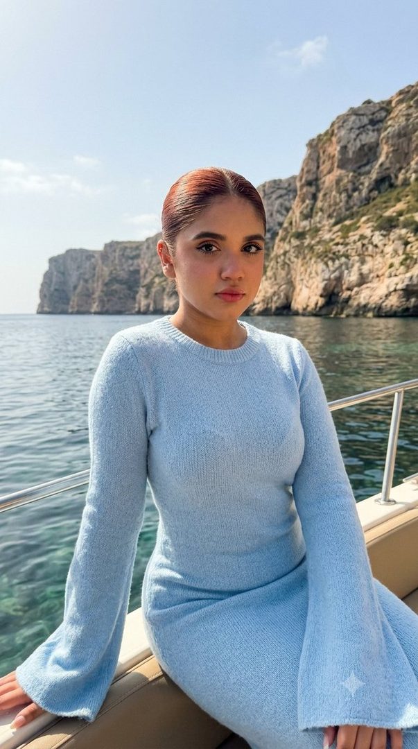 A young woman with reddish-brown hair pulled back, wearing a light blue knitted dress, sits on a boat with cliffs and the sea in the background.
