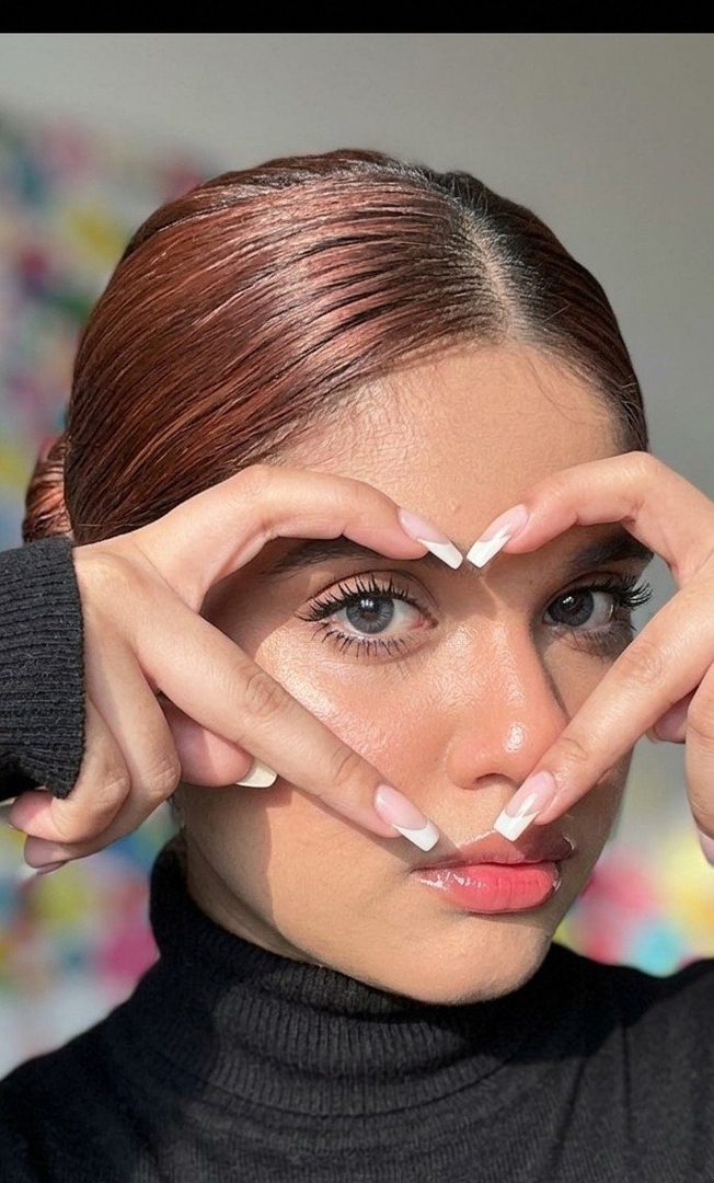 Close-up portrait of a young woman with brown hair, forming a heart shape with her hands around her eye, wearing a black turtleneck.