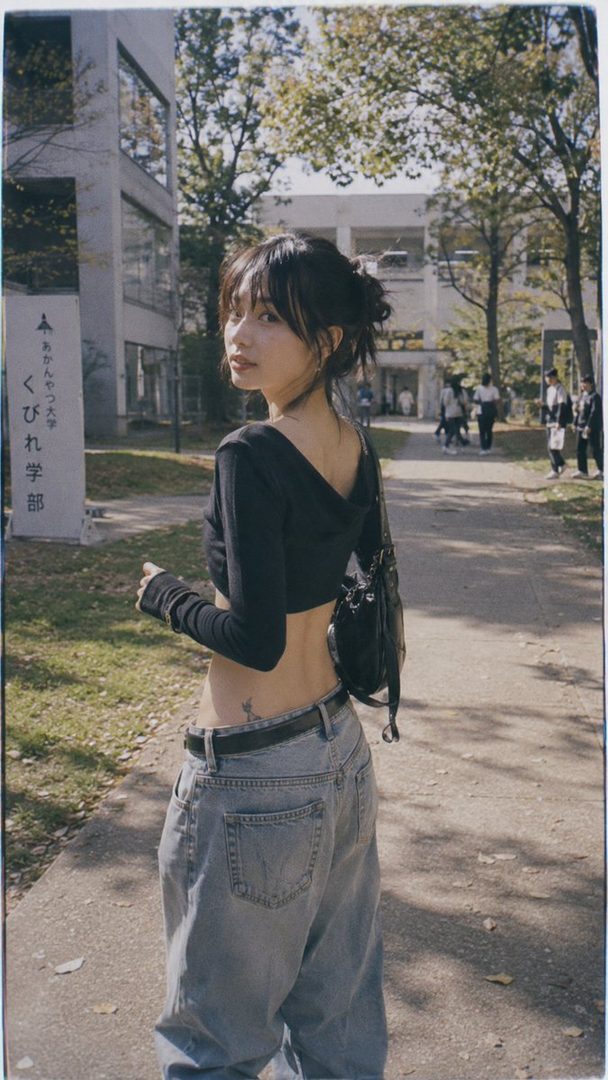 A young woman with her hair in a messy bun, wearing a black crop top and baggy jeans, stands on a campus path, looking over her shoulder.
