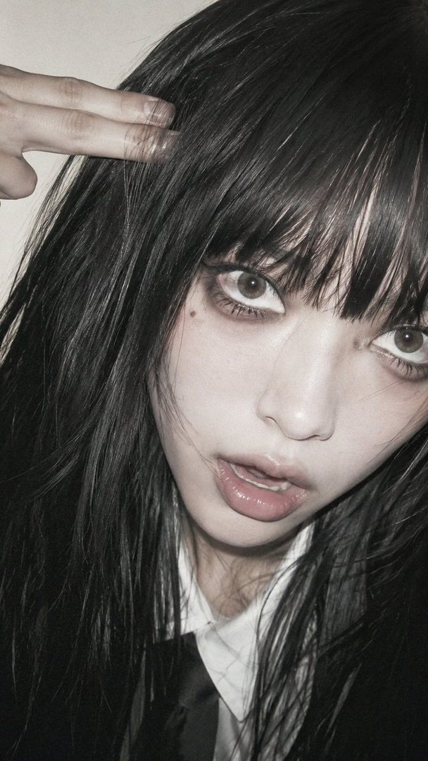 Close-up portrait of a young East Asian woman with long black hair and bangs, wearing a white blouse and tie, with heavy eye makeup and glossy lips.
