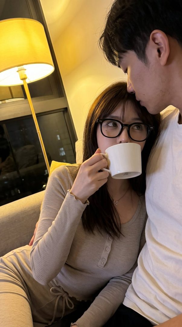 A couple cuddling on a couch at night, the woman drinks from a mug and wears glasses.