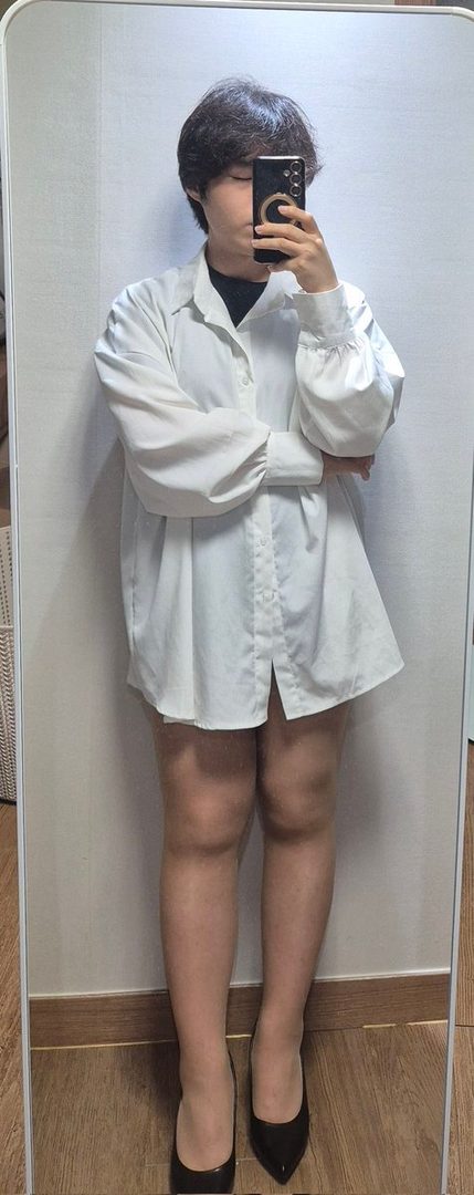 A person with short dark hair takes a mirror selfie, wearing an oversized white button-up shirt and black heels indoors.