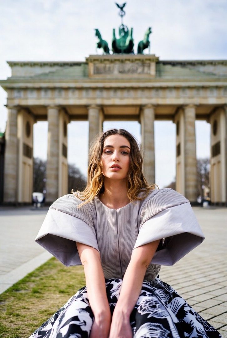 A woman with long blonde wavy hair sits in front of the Brandenburg Gate in Berlin, wearing a grey top with dramatic sleeves and a black and white patterned skirt.