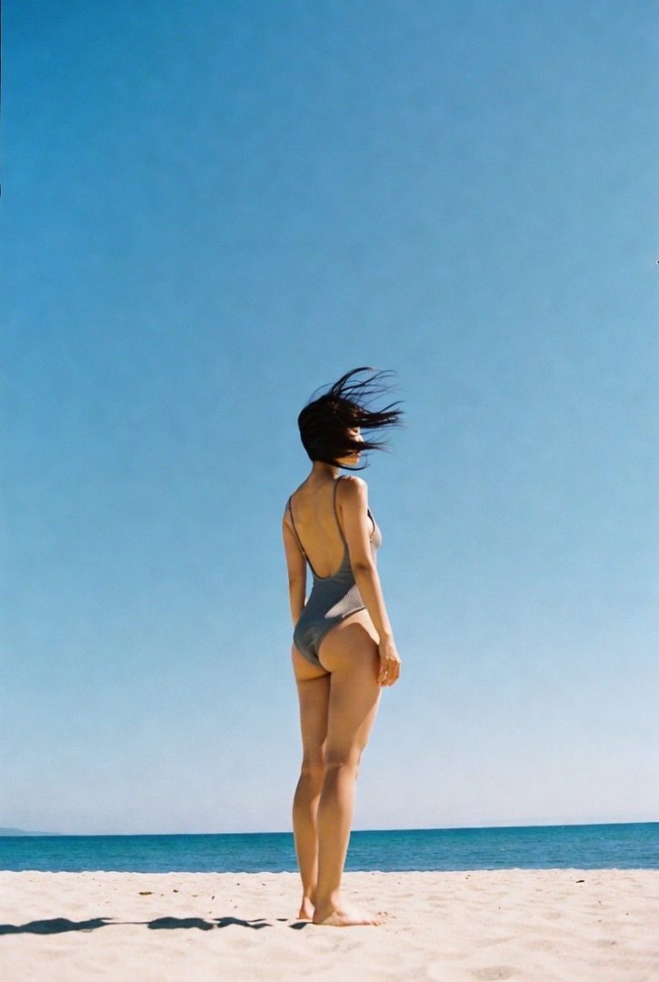A woman in a grey one-piece swimsuit stands on a sandy beach, looking out at the ocean with her hair blowing in the wind.
