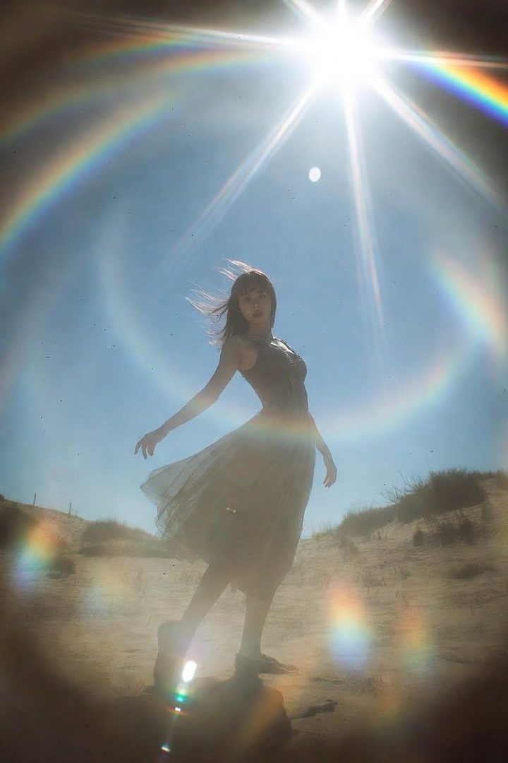 A young woman in a flowing dress stands in a sandy landscape under a bright sun with a rainbow halo effect.