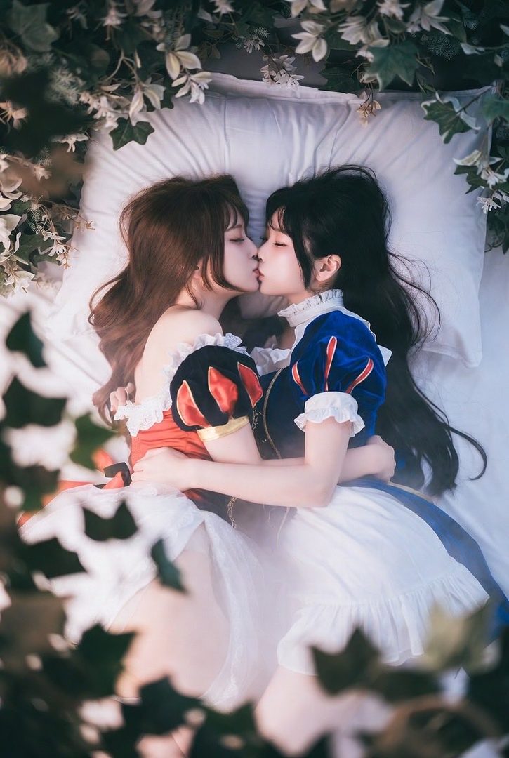 Two young women in fairytale-inspired dresses are lying down and kissing, surrounded by greenery.