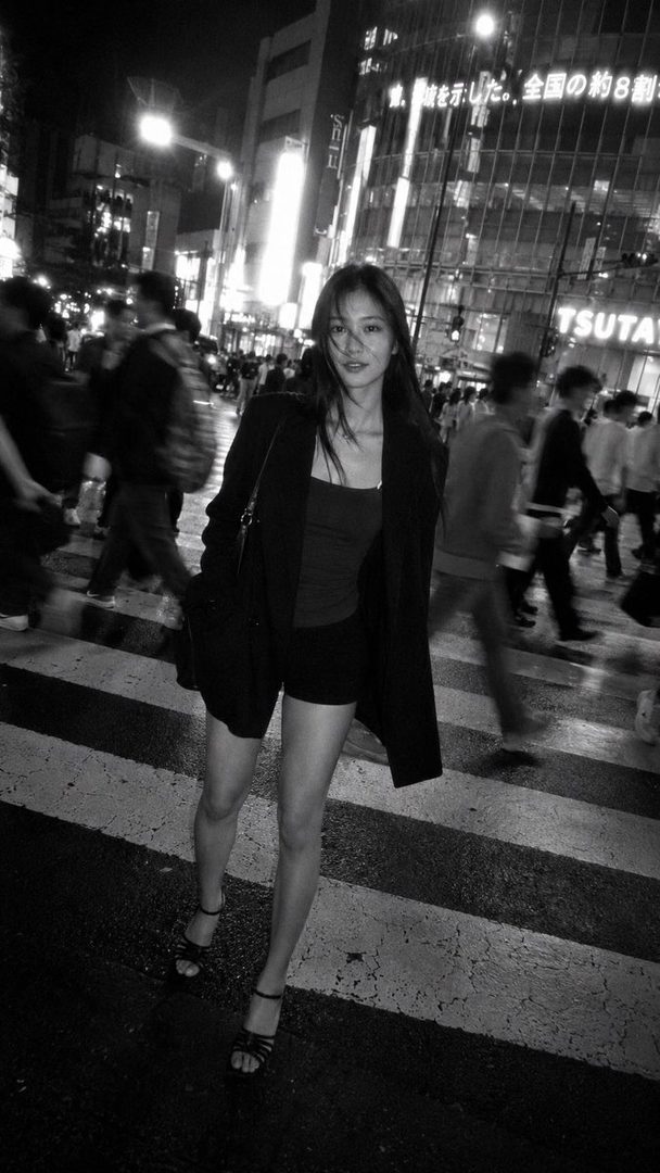 A young woman walks across a busy city street at night, wearing a blazer over a tank top and shorts.