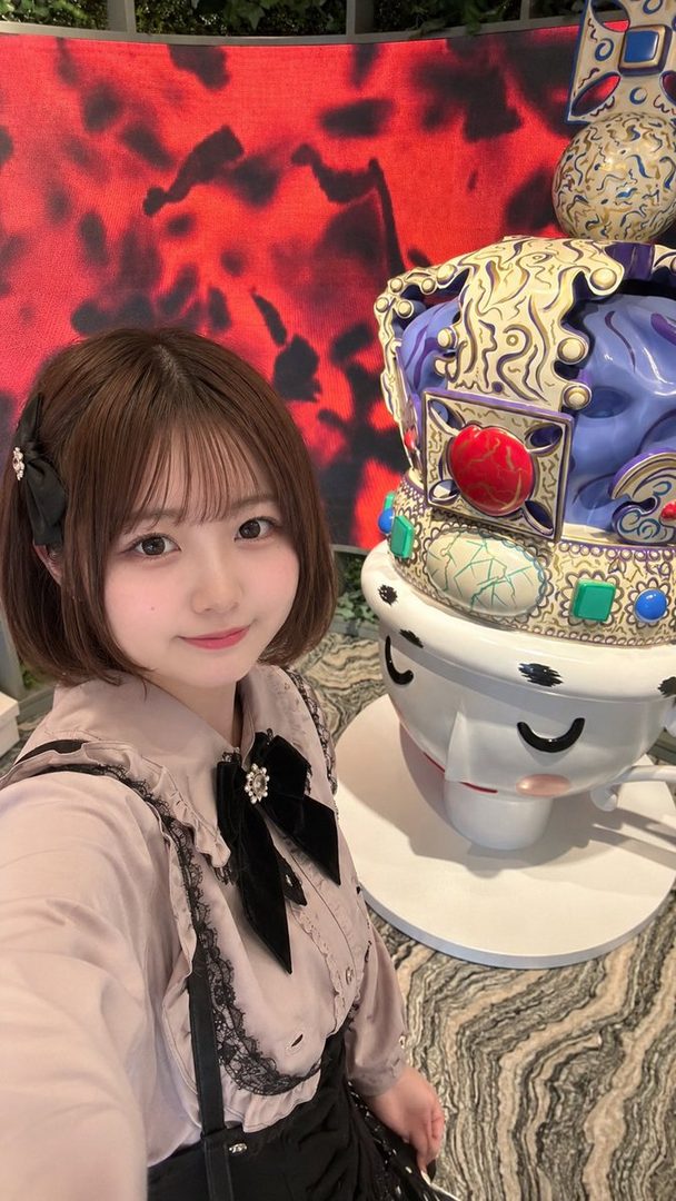 A young East Asian woman with short brown hair and bangs, wearing a light-colored blouse with black lace trim and a black bow, takes a selfie in front of a large, colorful, abstract sculpture.