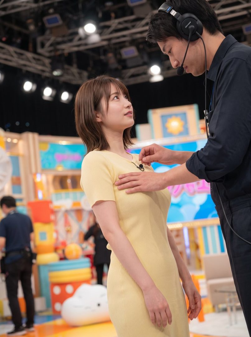 A TV crew member adjusts a microphone on a female news anchor in a bright studio setting.