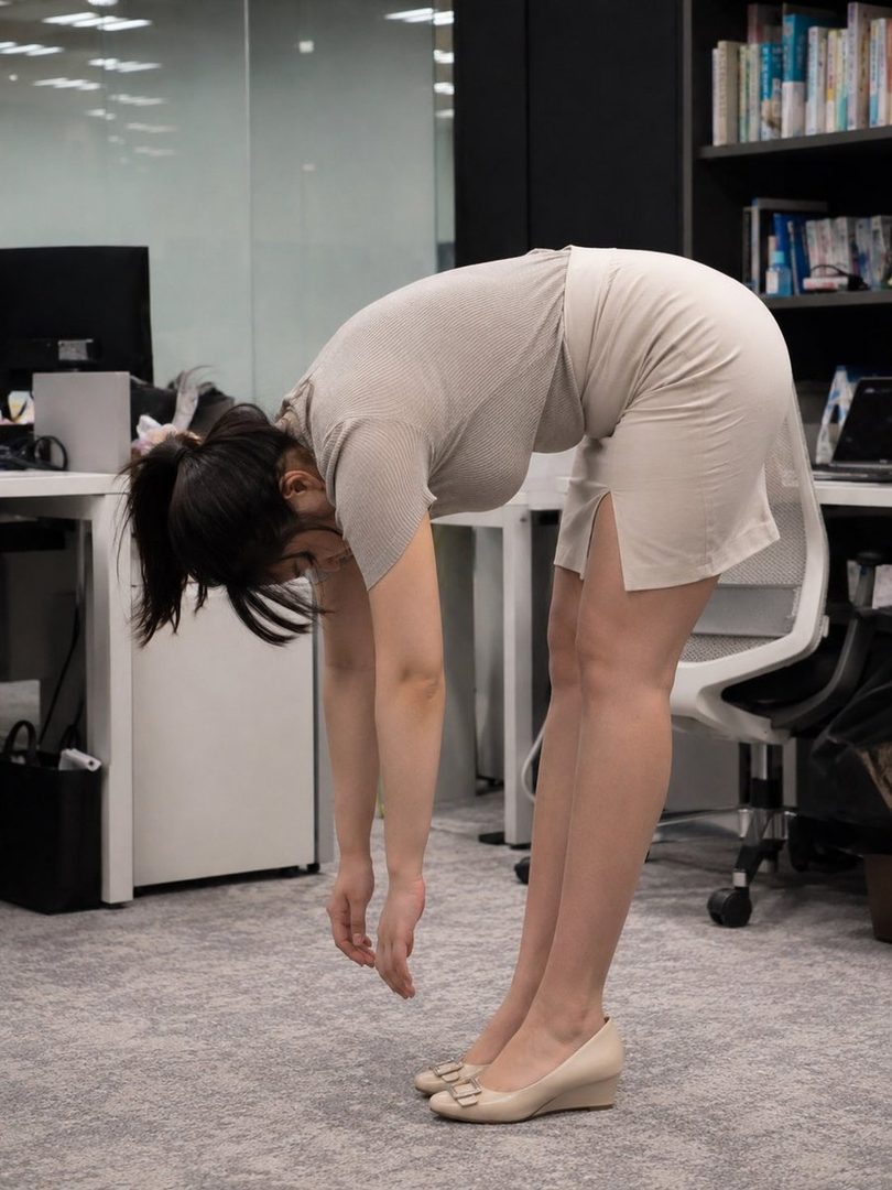 A woman in an office setting stretches by bending forward, her arms hanging loosely.